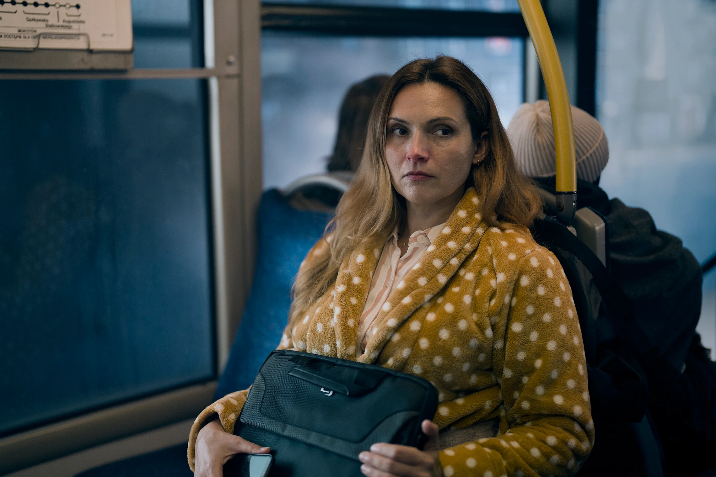Elegancka kobieta, z wyraźnymi oznakami przeziębienia, ubrana w brązy szlafrok w
kropki ściska teczkę i telefon w autobusie